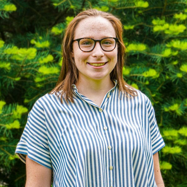 Headshot of Sarah in front of a green, pine tree wearing a button down striped shirt.