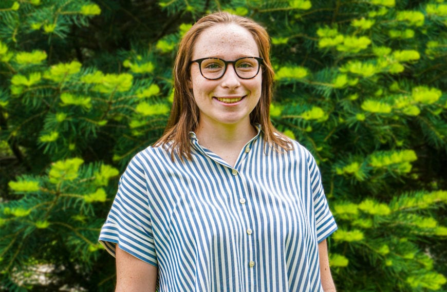 Headshot of Sarah in front of a green, pine tree wearing a button down striped shirt.