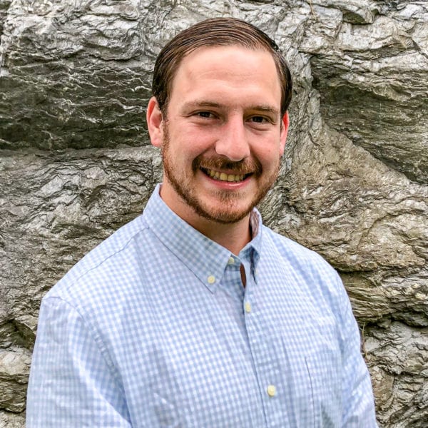 A photo of Joseph wearing a blue button-down shirt in front of grey-rock background/