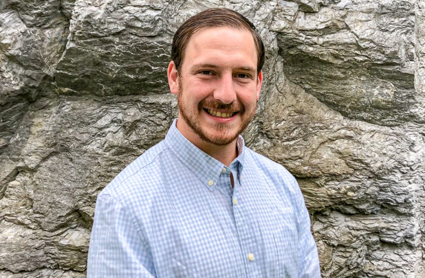 A photo of Joseph wearing a blue button-down shirt in front of grey-rock background/