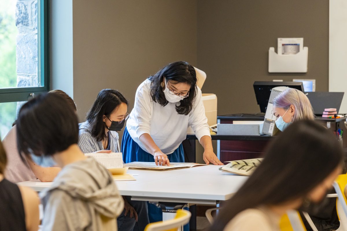 Kimiko Suzuki bends over a table in Lutnick Library going over documents with two masked students
