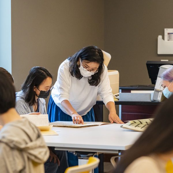 Kimiko Suzuki bends over a table in Lutnick Library going over documents with two masked students