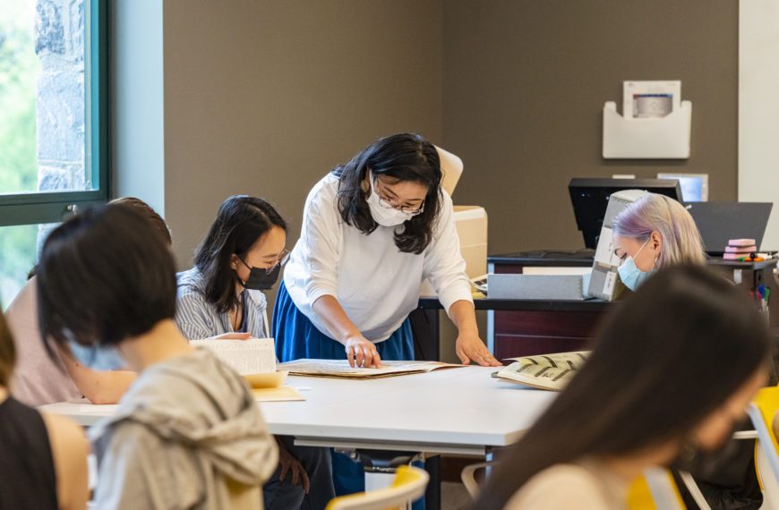 Kimiko Suzuki bends over a table in Lutnick Library going over documents with two masked students