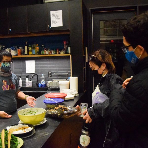 The professor and two students wear masks standing over bowls of food on the counter.