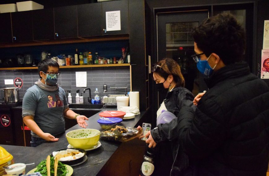 The professor and two students wear masks standing over bowls of food on the counter.