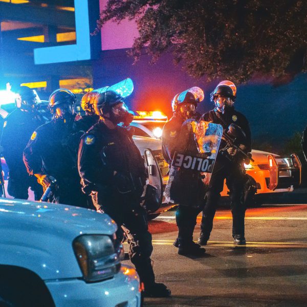Police gather in riot gear at night in front of a police car