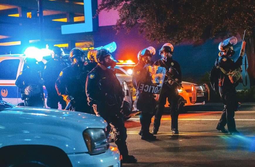 Police gather in riot gear at night in front of a police car