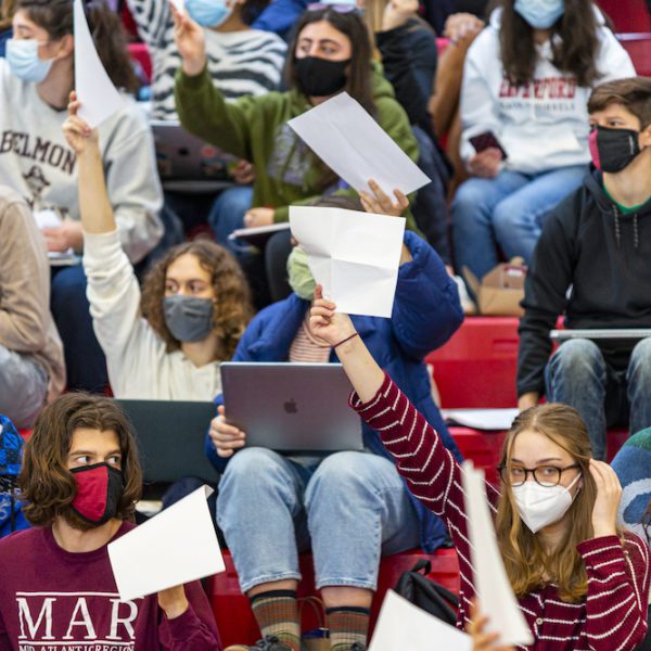 Masked students on bleachers in the basketball arena hold up white papers to signal their votes.