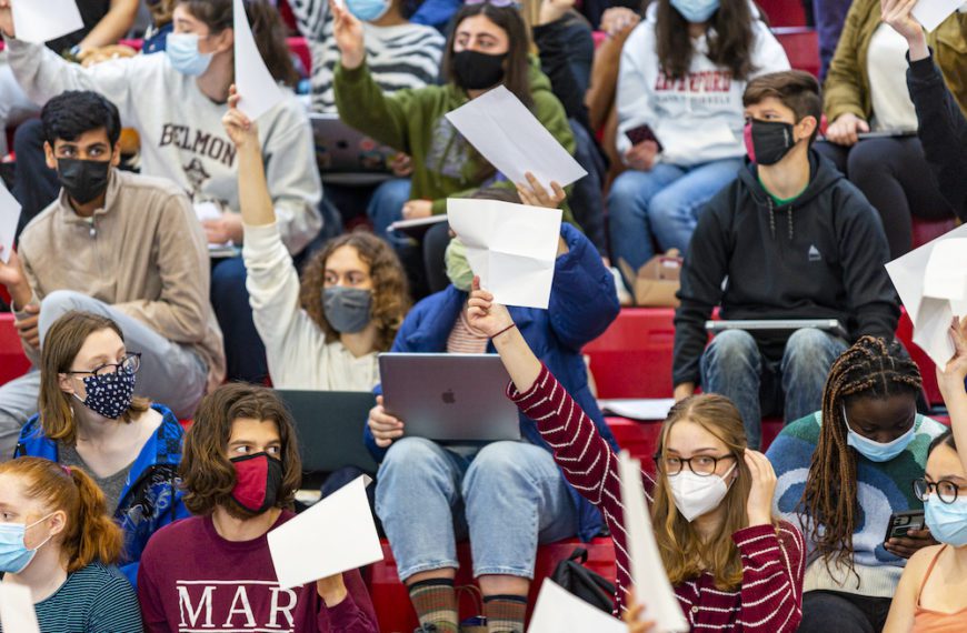 Masked students on bleachers in the basketball arena hold up white papers to signal their votes.