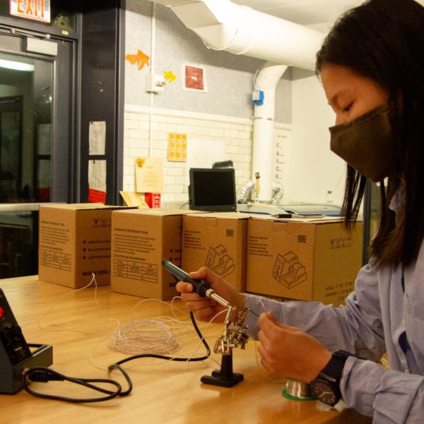 A masked student works on soldering in at a long table in the Maker Arts Space.