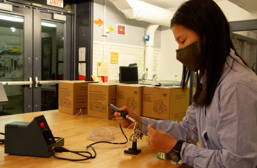 A masked student works on soldering in at a long table in the Maker Arts Space.