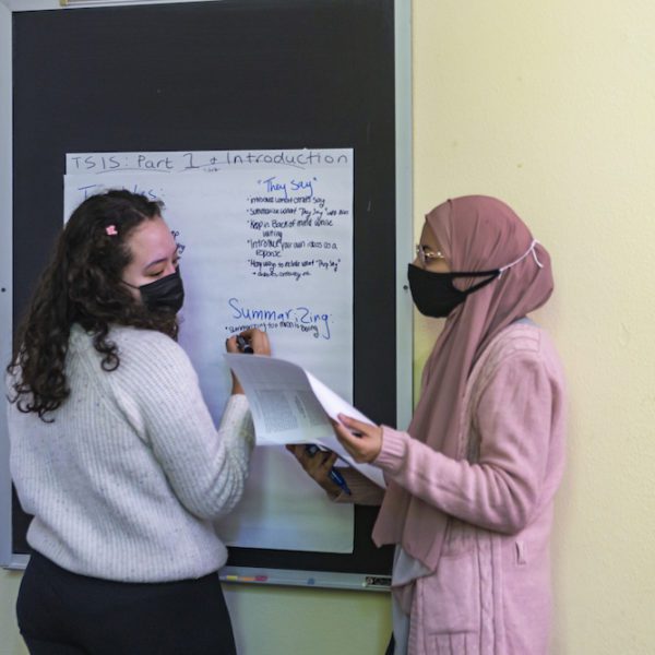 Two students, both wearing masks, write on a large piece of paper tacked up to the chalkboard. Categories for "They Say" and "Summarizing" can be seen.