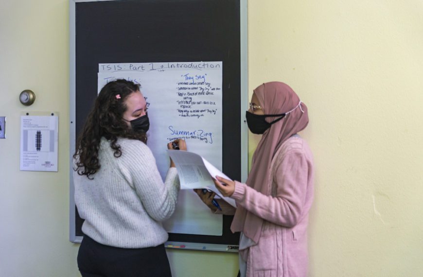 Two students, both wearing masks, write on a large piece of paper tacked up to the chalkboard. Categories for "They Say" and "Summarizing" can be seen.