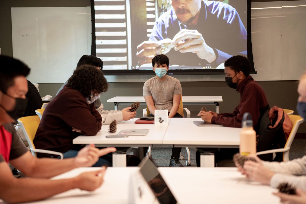 Students wearing masks surround the masked professor, who sits under a projection screen, as they pass around a pine cone.