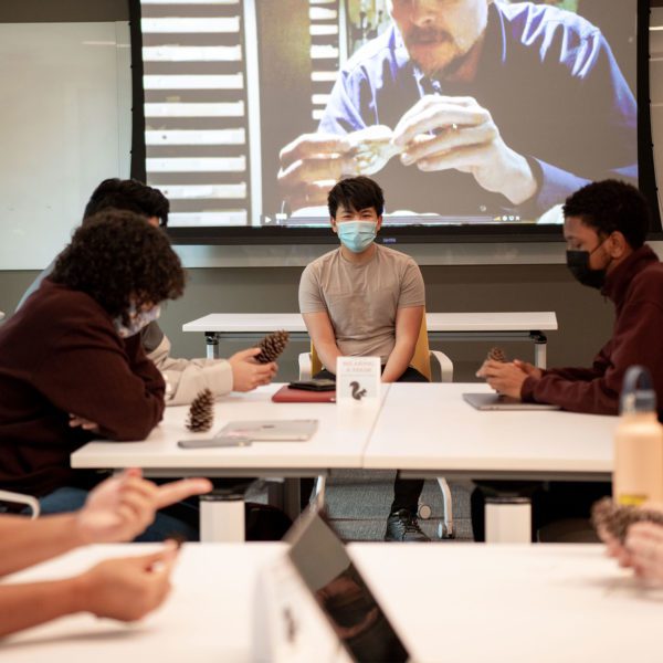 Students wearing masks surround the masked professor, who sits under a projection screen, as they pass around a pine cone.