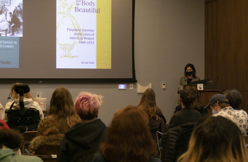 Jess Libow wears a mask and speaks at a podium in front of a projection screen with photos of the covers of the books "The Eternally Wounded Woman" and "Physical Culture and the Body Beautiful" on it.