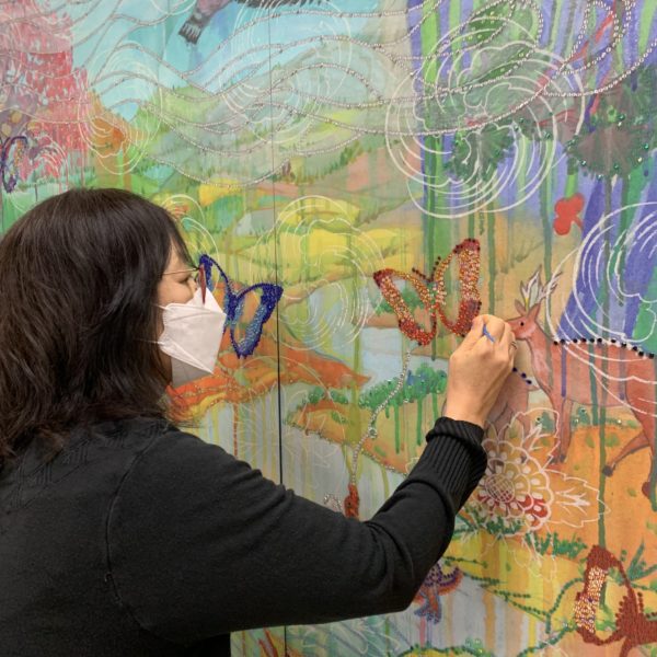 The artist, wearing a mask, applies red rhinestones to a butterfly in the painting.