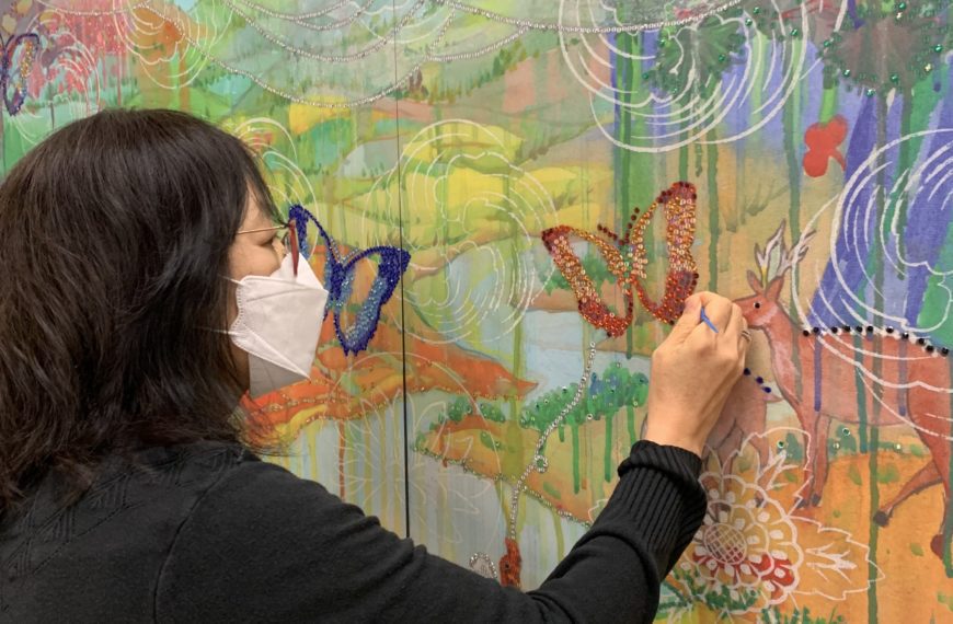 The artist, wearing a mask, applies red rhinestones to a butterfly in the painting.