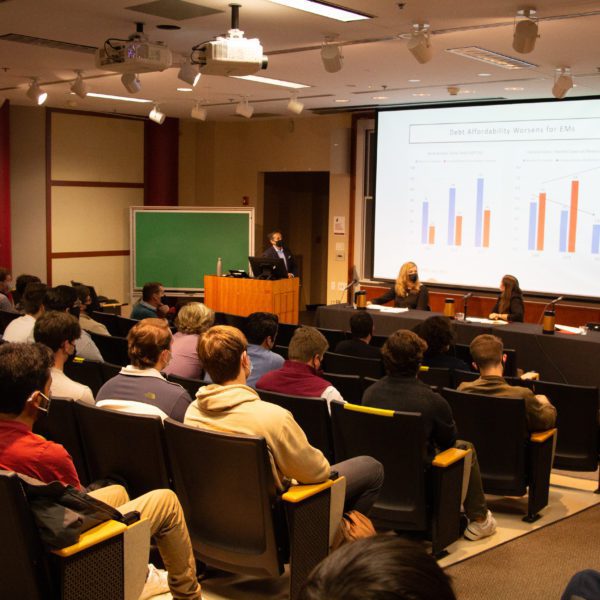 A speaker at the podium while three other alumni sit under a screen with a large graph on it depicting "an elusive hunt for credit convergence"