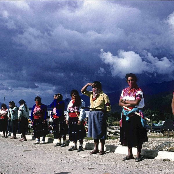 Women with scarves over their noses and mouths stand in a line on the side of the road with blue skies and mountains behind them.