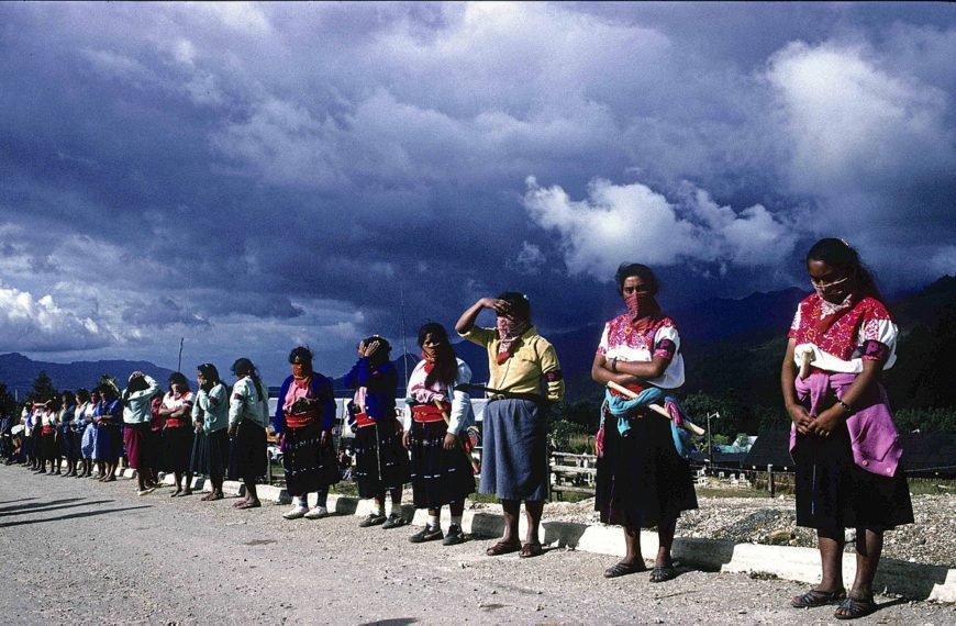 Women with scarves over their noses and mouths stand in a line on the side of the road with blue skies and mountains behind them.