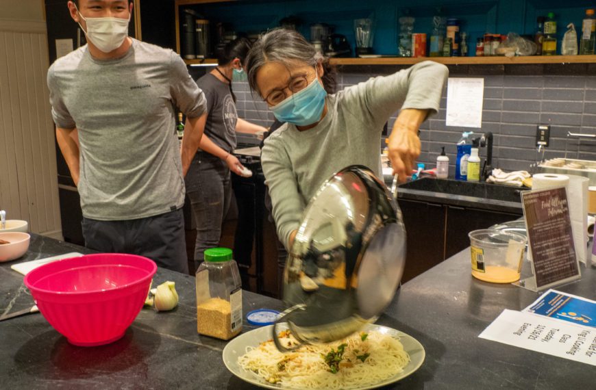 Ying Li wears a mask and pours noodles out of a wok as a masked student stands behind her in the VCAM community kitchen.