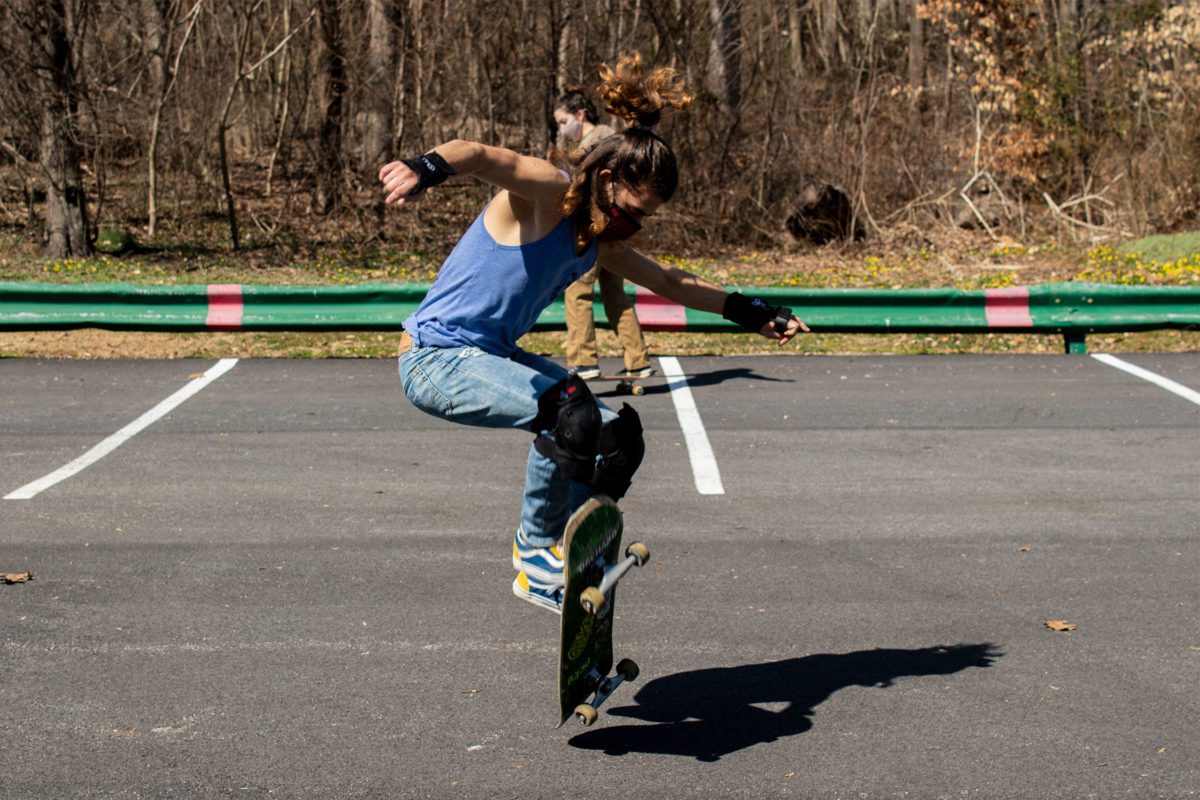 A student wearing a mask, tank top, and jeans, ollies on a skateboard in the parking lot.
