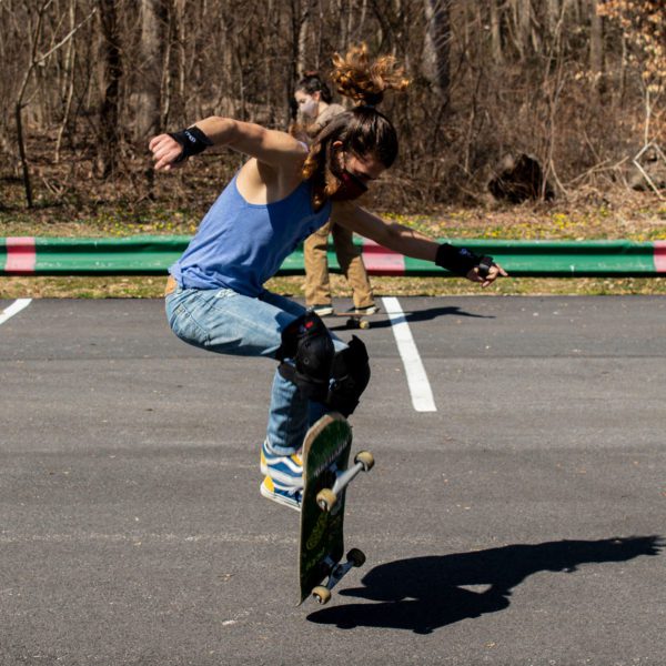 A student wearing a mask, tank top, and jeans, ollies on a skateboard in the parking lot.