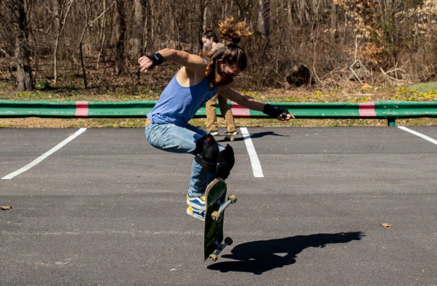 A student wearing a mask, tank top, and jeans, ollies on a skateboard in the parking lot.