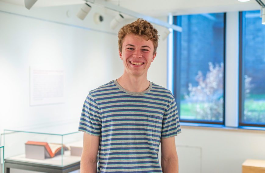 Nick wears a striped shirt and smiles in the gallery in the library.