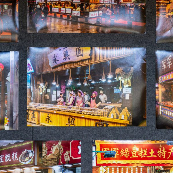 Seven photos of Chinese street food in the exhibit, and in the center a photo of four women with purple scarves on their heads standing behind a counter serving food.