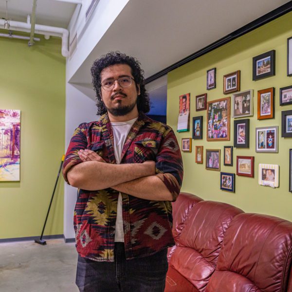Henry stands in front of a red vinyl couch and a wall of photographs with one of his paintings on the opposite walls.