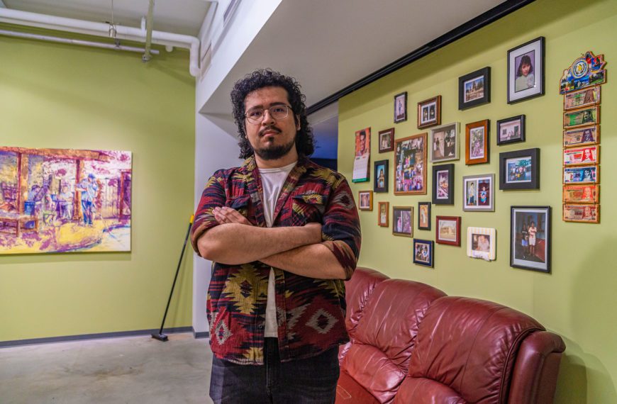 Henry stands in front of a red vinyl couch and a wall of photographs with one of his paintings on the opposite walls.