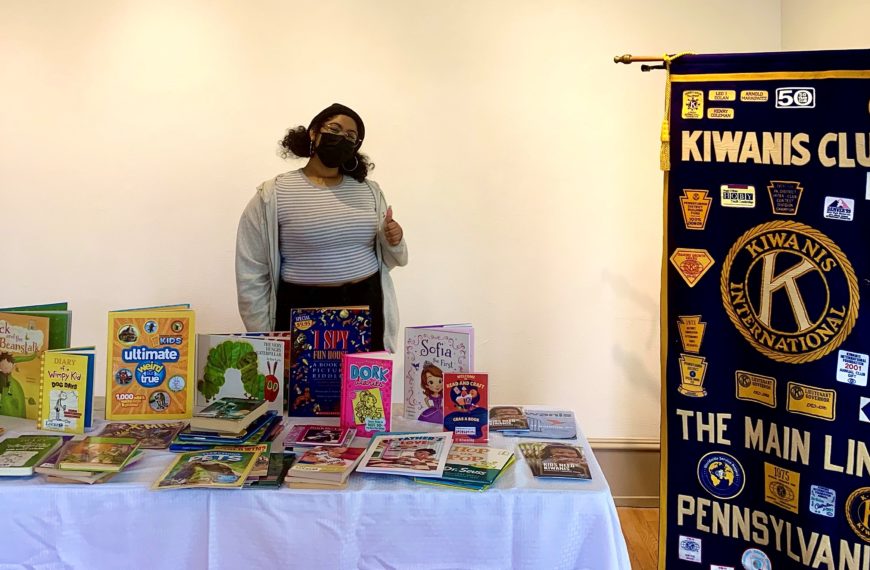 Jalexie stands next to a Kiwanis Club banner and behind a table full of children's book.