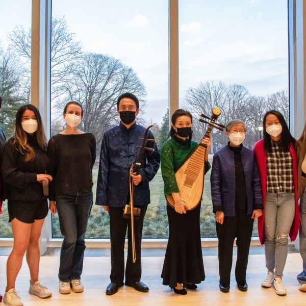 Eight masked people stand in front of a wall of windows in the concert hall, two of whom are holding a erhu (a two-stringed fiddle) and pipa (lute).