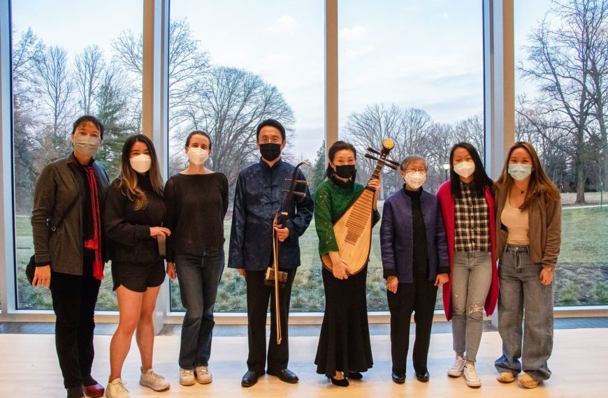 Eight masked people stand in front of a wall of windows in the concert hall, two of whom are holding a erhu (a two-stringed fiddle) and pipa (lute).