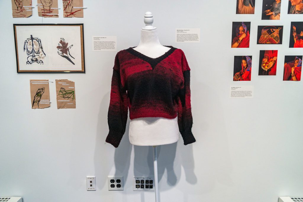 An installation of anatomy drawings with thread embroidery through them next to a black and red sweater on a dressform.