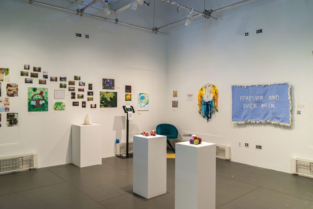 Three stands with sculptures on them and two walls covered in fabric art and many different sizes and styles of drawings and paintings.