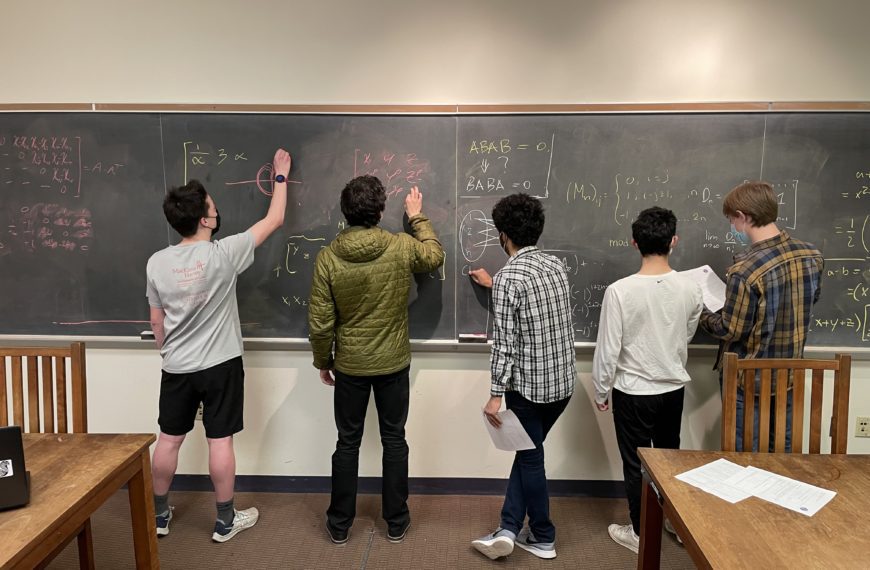 Five students stand in front of a chalkboard working on math problems together.