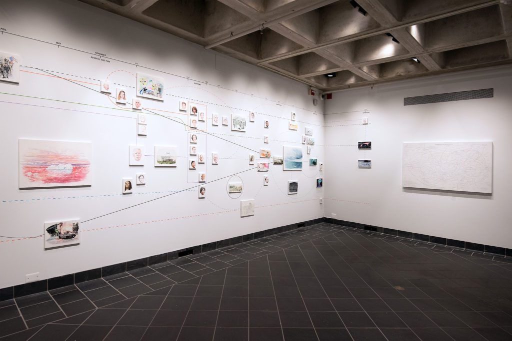 Several drawings, including one of an iceberg against a pink sunset and several portraits including a screaming Trump, Amy Coney Barrett, and Joe Biden, connected by lines on the gallery wall.