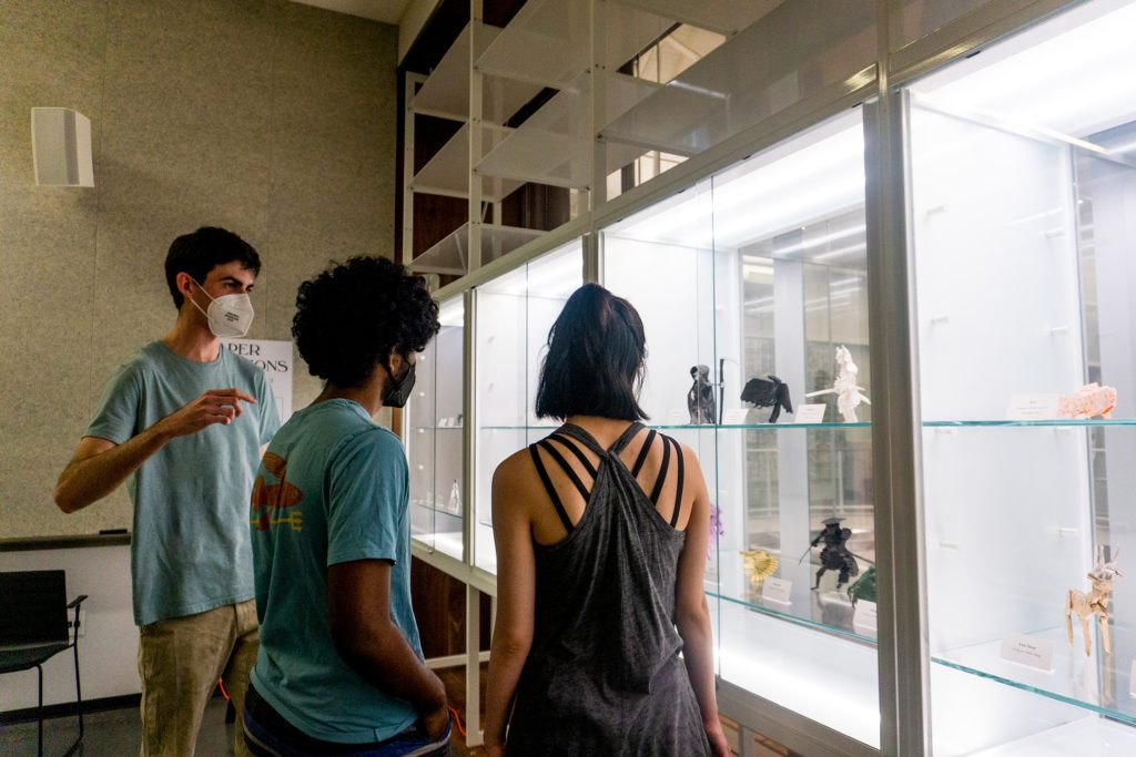 Chris, wearing a mask, explains his work to two students as they all stand in front of the display case.