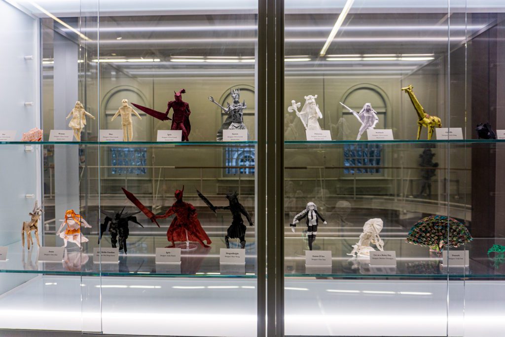 A display case with 16 different colorful origami pieces in it, including a peacock, a giraffe, and several warriors with swords.