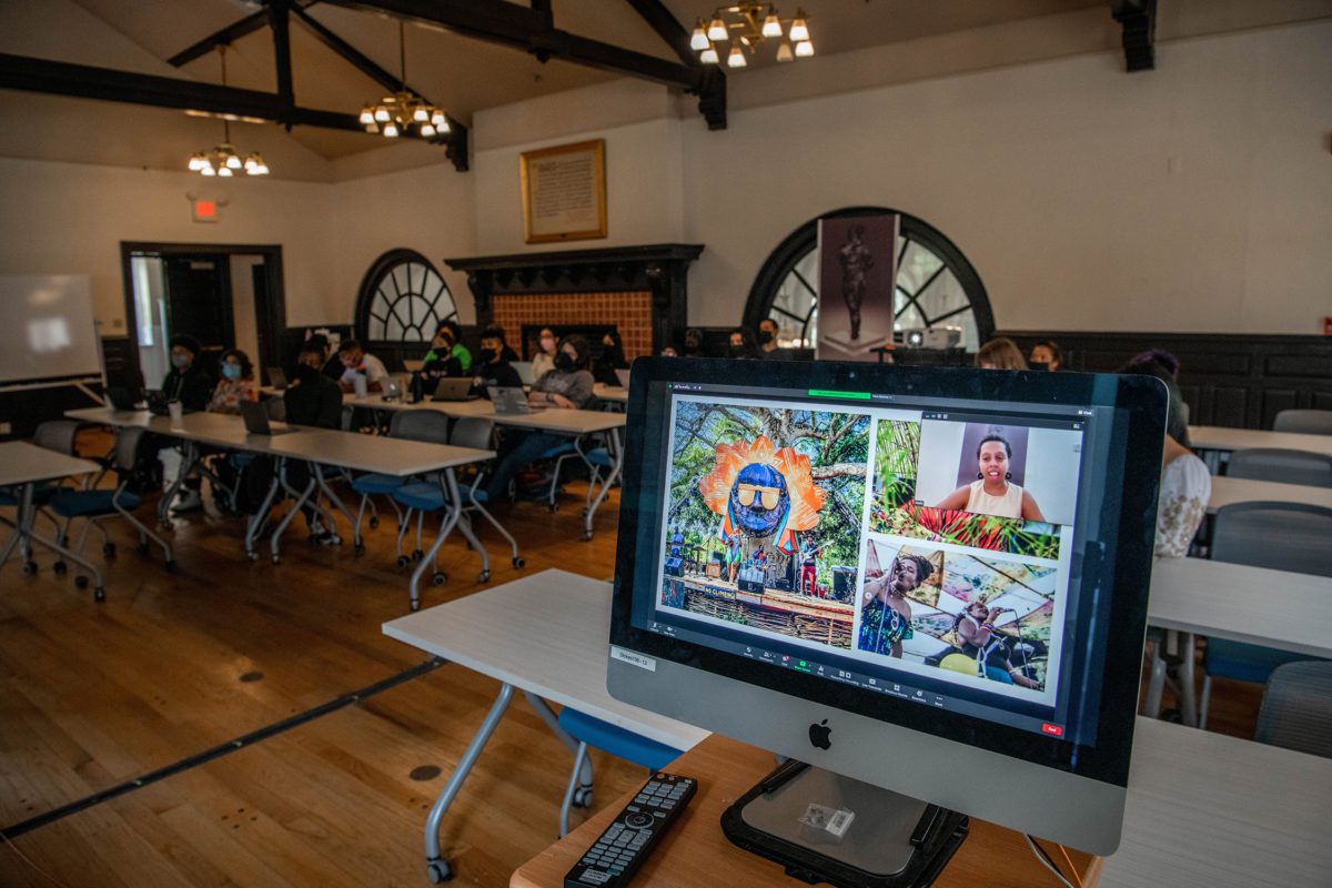 A computer screen featuring the artist live on Zoom and some of their colorful works on display--including one of a live band performing--with the seated class in the background.