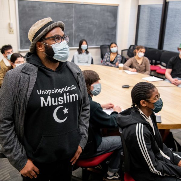 The class sits around a table with Guangtian standing on the far left and Musa standing in the foreground wearing a sweatshirt that says "Unapologetically Muslim"
