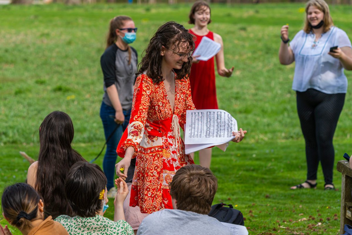 A student in a sundress holds a musical score in one hand and hands another student a flower with another.