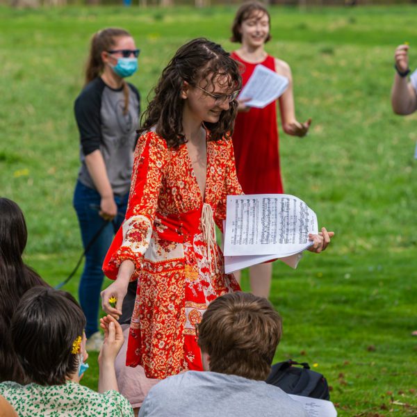 A student in a sundress holds a musical score in one hand and hands another student a flower with another.