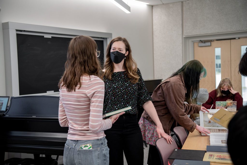 Rachel wears a mask and talks to a student next to a piano while other students look through books set out on the table.