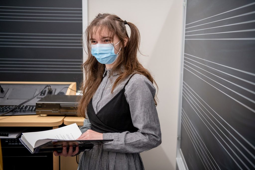 A student in pigtails and a mask holds a score and stands in front of a blackboard with musical staffs on them.