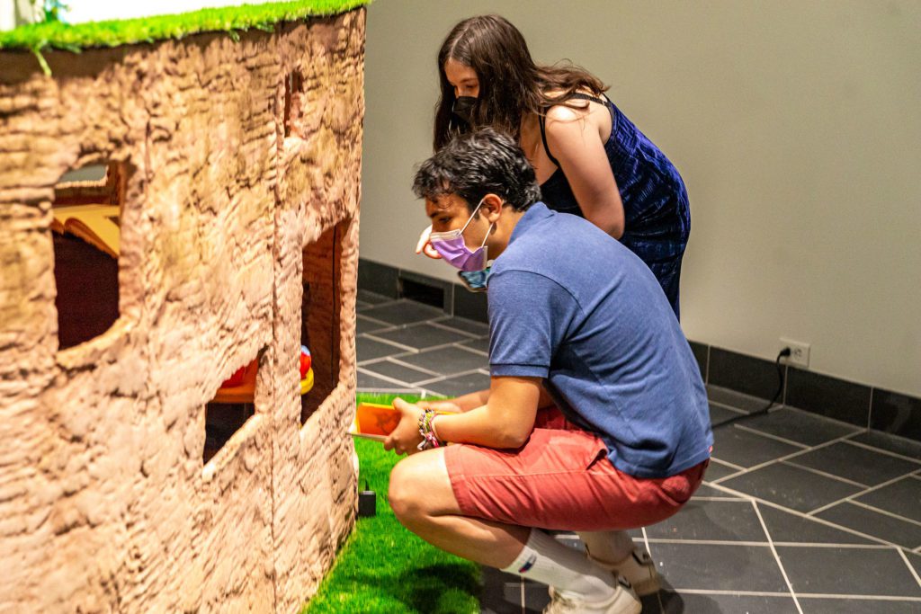 Two masked students crouch next to the air-dried clay structure.