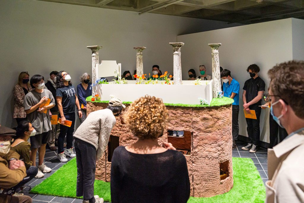 A crowd gathers around a large installation that includes an air-dried clay structure with plants growing on it.